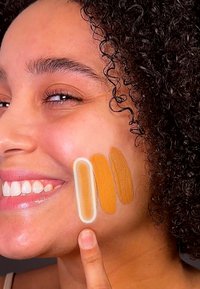 Fondasjonsprøver i tre nyanser på kinnet, med en glatt, matt finish i middels til dype gyldne toner. Hudteksturen er synlig.