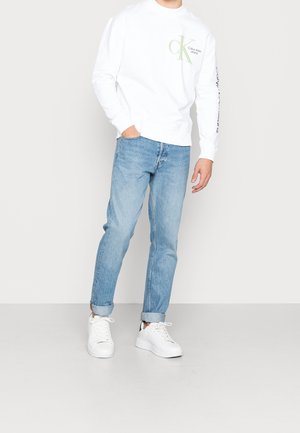 Felpa bianca in cotone con logo verde, jeans in denim blu e sneakers bianche. Vestibilità casual, con risvolti sui jeans e design minimale.
