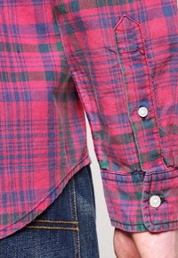 Czerwona i granatowa koszula w kratę z rękawem i zapięciem na guzik, z lekkim połyskiem, widocznymi detalami szwów i zaokrąglonym dołem. Noszona na dżinsy.