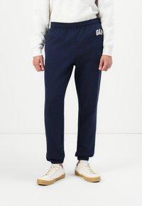 Marineblå joggebukser med elastisk livlinje, ribbede mansjetter og "GAP"-logoet brodert på venstre hofte. Myk tekstur.