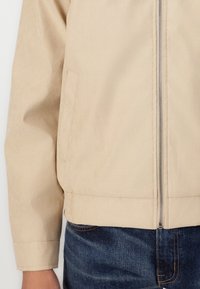 Veste zippée beige en tissu lisse, dotée de poches latérales et d'une taille ajustée, associée à un jean en denim bleu foncé.