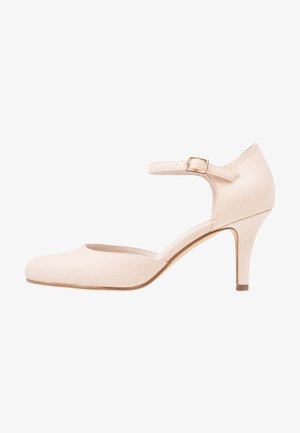 Beige högklackad sko med en slät ovandel och texturerad detaljering. Har en justerbar vristrem och en smal klack.