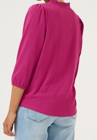 Blouse fuchsia avec une finition texturée, dotée de manches bouffantes trois-quarts et d'un col à volants. Portée avec un jean bleu clair.