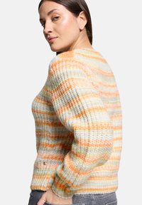 Kobieta z ciemnymi włosami, ubrana w gruby sweter typu oversize w paski w kolorze pomarańczowym, żółtym i szarym, spogląda przez ramię na jednolitym tle.