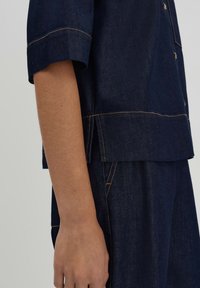 Dark denim shirt featuring a straight cut, short sleeves, and contrasting orange stitching along the hems and seams. Visible side slit detail.