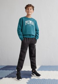 Sudadera de color azulado con el logotipo "PUMA" en blanco y pantalones de chándal negros con logotipo, combinados con zapatillas deportivas negras. Fondo: baldosas de suelo texturizadas.