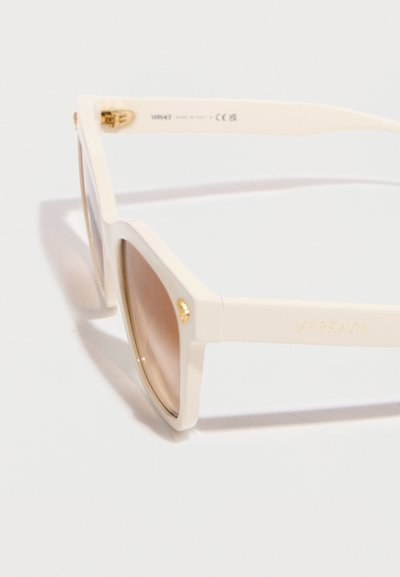 Lunettes de soleil Versace couleur crème avec montures carrées, verres dégradés marron, et emblème doré de Méduse sur les branches et les coins des montures.
