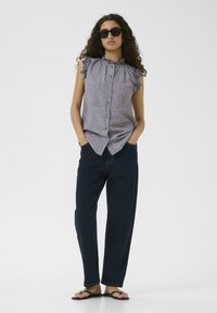 Hallhallik koduõhuga ilma varrukatega pluus, millel on volangid, nuppude esiküljel, koos siniste laiad jalad teksade ja mustade sandaalidega valge tausta ees.