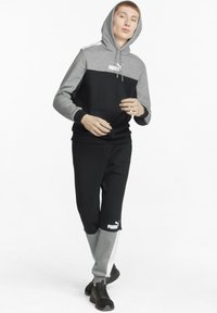 Sudadera con capucha gris y negra con un bolsillo tipo canguro y un logo; combinada con pantalones negros con detalles grises y un logo en la pierna.