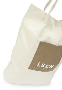Tasche aus cremefarbenem Stoff mit einer rechteckigen braunen Tasche, die weißen Text enthält. Einfaches Design mit zwei Traggriffen.