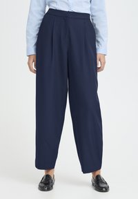 Tumesinised püksid kõrge vöökohaga, millel on voldid ja mugav lõige. Valmistatud siledast kangast, kantud mustade mokassiinidega.