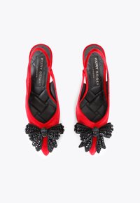 Czerwone zamszowe buty na obcasie mają czarną koralikową kokardę na palcach, pikowaną czarną wyściółkę wewnętrzną oraz otwartą piętę.