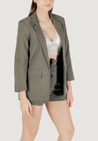 Blazer verde oliva con una consistenza liscia, caratterizzato da un bottone singolo e due tasche frontali, indossato sopra un reggiseno in pizzo bianco e pantaloni shorts.