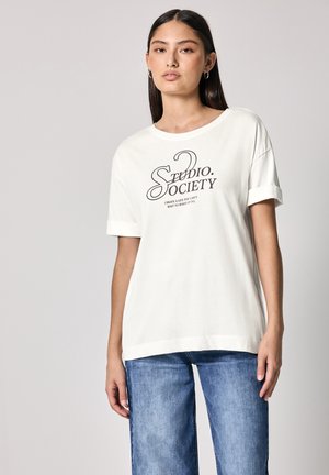 T-shirt en coton biologique écru avec des manches retroussées, comportant un texte imprimé noir "Studio Society" et une coupe décontractée. Jeans en bleu clair.