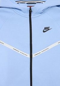 Svijetloplava zip-up majica sa kapuljačom, s crnim patentnim zatvaračem, Nike logotipom i bijelim naglaskom "NIKE TECH" duž prsnog dijela u geometrijskom dizajnu.