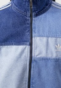 Jeansjacka med dragkedja framtill, med färgblockdesign i mörka och ljusa blå nyanser, slät textur och broderad logotyp.