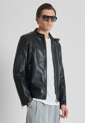 Man draagt een zwarte leren jas, wit T-shirt, grijze broek en donkere zonnebril tegen een effen lichtgrijze achtergrond.