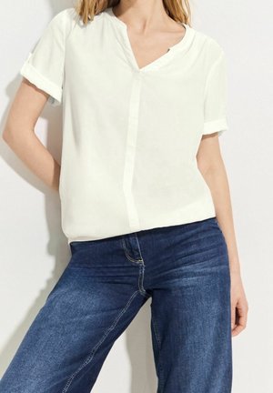 Woman wearing a short-sleeve white blouse and blue jeans, standing with one hand on hip against a plain light background.