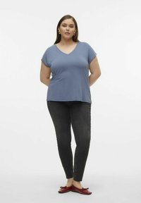 Modrý v krku tričko z měkké látky, spárované s tmavě šedými slim-fit džíny a červenými plochými botami. Modelka stojí s rukama na bocích.