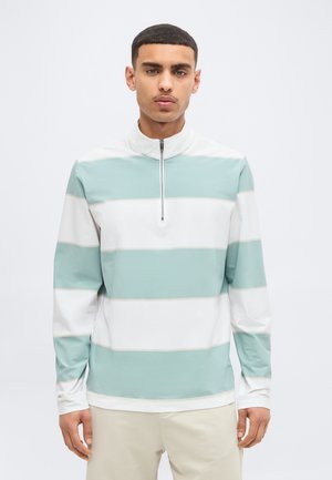 Άνδρας που φοράει πουλόβερ με μακριά μανίκια και μισό φερμουάρ, με φαρδιές οριζόντιες ρίγες σε λευκό και ανοιχτό πράσινο, στέκεται μπροστά σε απλό φόντο.