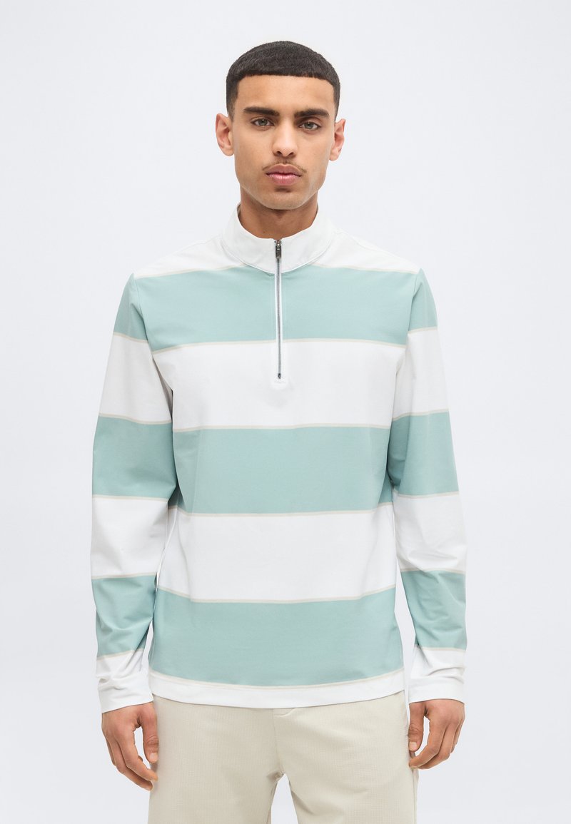 Homme portant un pull à manches longues, à fermeture éclair sur la moitié du devant, avec de larges rayures horizontales blanches et vert clair, se tenant devant un fond uni.