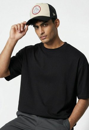 Junger Mann, der ein schwarzes T-Shirt und graue Hose trägt, richtet eine beige und schwarze Baseballkappe mit Berglogo vorne.