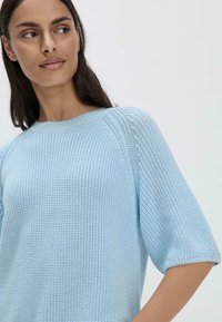 Vaaleansininen neulepaita, jossa on pyöreä kaula-aukko ja lyhyet puffihihat. Siinä on tekstuurinen kuvio ja ribbaushelma.