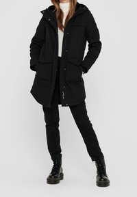 Parka negra impermeable con capucha, dos bolsillos frontales y cremallera completa. Llevada sobre una camiseta blanca y combinada con pantalones negros y botas.
