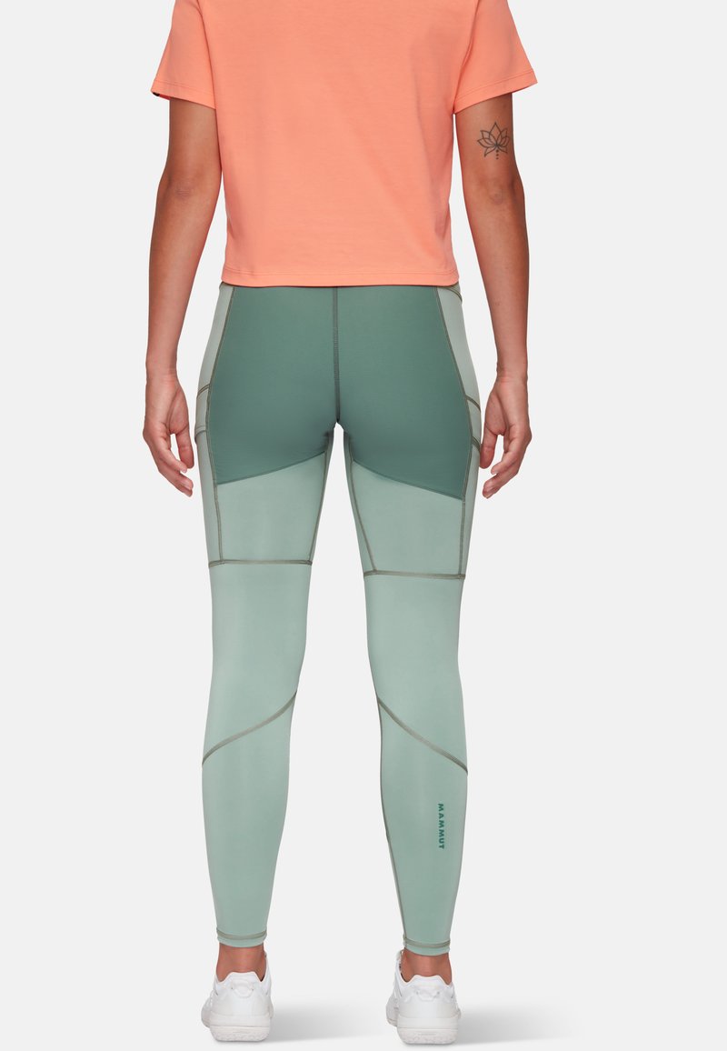 Person, die mintgrüne und dunkelgrüne, color-blocked Leggings, ein korallenfarbenes, kurzes Oberteil, weiße Sneaker und ein Lotus-Tattoo am linken Arm trägt.