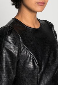 Svart lädertop med strukturerad yta, som har samlade puffärmar och rund halsringning. Slät yta med ett subtilt mönster.