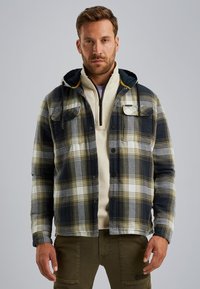 Chaqueta de franela a cuadros en navy, blanco y verde con capucha, que cuenta con dos bolsillos en el pecho y una textura suave, sobre una sudadera crema con cremallera.