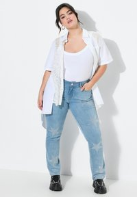 Camisa blanca de manga corta sobre una blusa blanca, combinada con jeans de mezclilla azul claro con adornos de estrellas plateadas. Zapatos negros de suela gruesa.