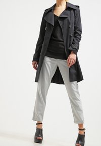 Czarny trencz z matowym wykończeniem, szerokim kołnierzem i detalami z guzikami, zestawiony z szarymi, dopasowanymi spodniami oraz sandałami na masywnym obcasie.