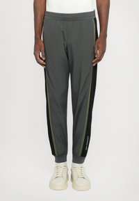 Pantalons de sport grays avec une taille élastique, des rayures latérales noires et vert olive contrastantes, et des poignets fuselés. Portés avec des baskets de couleur claire.