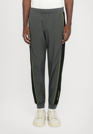Pantalons de sport grays avec une taille élastique, des rayures latérales noires et vert olive contrastantes, et des poignets fuselés. Portés avec des baskets de couleur claire.