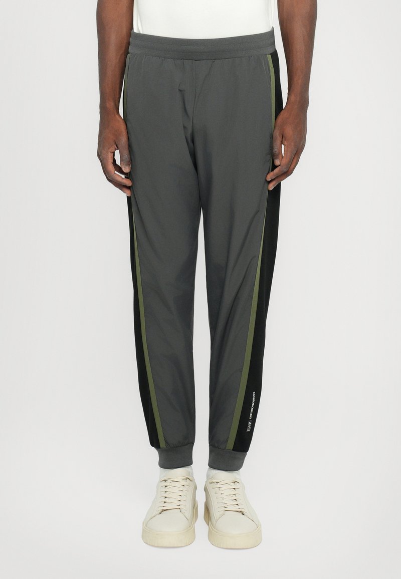 Pantalons de sport grays avec une taille élastique, des rayures latérales noires et vert olive contrastantes, et des poignets fuselés. Portés avec des baskets de couleur claire.