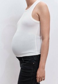 Persona embarazada vistiendo una blusa sin mangas blanca y pantalones negros, mostrada de perfil contra un fondo simple.