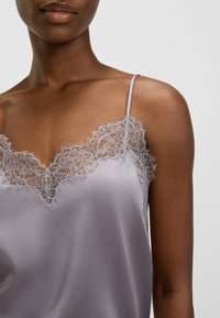 Eine dunkelhäutige Person trägt ein lilafarbenes Satin-Camisole mit zarter, floralem Spitzenrand und schmalen Trägern vor einem schlichten Hintergrund.