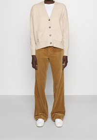 Beige pletený cardigan s véčkovým výstřihem, zapínáním na knoflíky a kapsami. Široké manžetové kalhoty z manšestru v hnědé barvě. Bílé tenisky dokončují tento outfit.