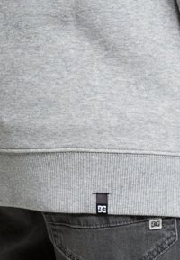 Sudadera gris con ribetes en el dobladillo y las mangas. Incluye una pequeña etiqueta negra de DC en la costura inferior. La tela parece suave y texturizada.