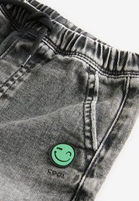 Grå denimshorts med delvis elastisk midja, med en grön smileyknappdetalj och synlig sömnad.