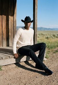 Homme portant un chapeau de cowboy noir, un pull beige et un pantalon noir assis sur une véranda en bois avec de l'herbe sèche et des montagnes lointaines en arrière-plan.