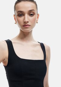 Vestido negro sin mangas con escote cuadrado, silueta ajustada y textura suave. Pendientes de aro dorados añaden un acento metálico.