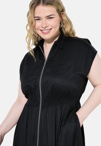 Abito nero con zip frontale e maniche corte, dotato di un elastico in vita e due tasche anteriori. Tessuto morbido con un design di colletto classico.