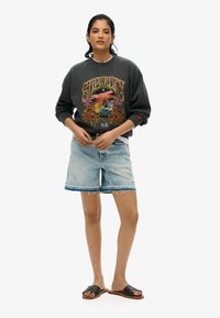 Vrouw in een donkere graphic sweatshirt, lichte blauwe denim shorts en zwarte slidesandalen, staand tegen een witte achtergrond.