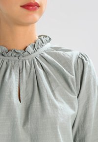Light green blouse with a ruffled neckline, keyhole opening, and gathered texture. Made from a soft, slightly shiny fabric.