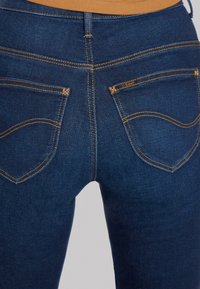 Mörkblå denimjeans med en åtsittande design, med orange sömmar och två bakfickor med en liten etikett på högra fickan.