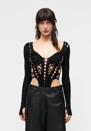 Γυναίκα με μακριά σκουρόχρωμα μαλλιά, που φορεί μαύρο μακρυμάνικο τοπ με μοτίβα κοψιμάτων και μαύρο δερμάτινο παντελόνι, στέκεται μπροστά σε σκέτο φόντο.