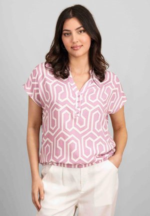 Rosa Bluse mit kurzen Ärmeln, die ein geometrisches weißes Muster zeigt, mit einem knöpfbaren Kragen und einem entspannten Schnitt, kombiniert mit cremefarbenen Hosen.