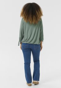 Zelený volný top s rukávy 3/4 a zavázanými manžetami, kombinovaný s modrými rozšířenými džíny s našitými zadními kapsami a béžovými botami.